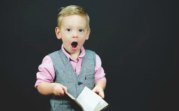 驚いた表情の幼い男の子が、開いた本を持ち、グレーのベストとピンクのシャツを着用しているポートレート。